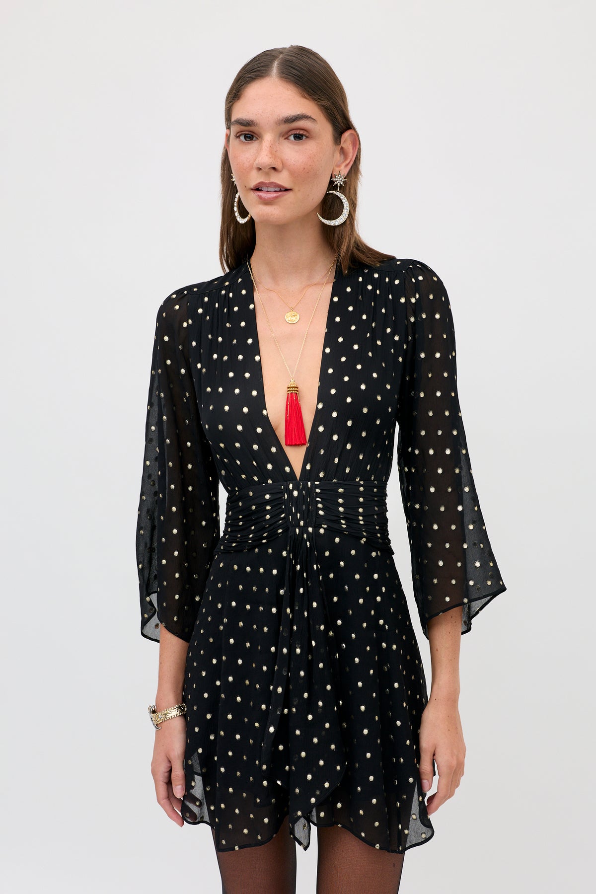 A woman wearing a Clove - Gold Spot Black long-sleeved mini dress, gold jewelry, and a red tassel necklace.