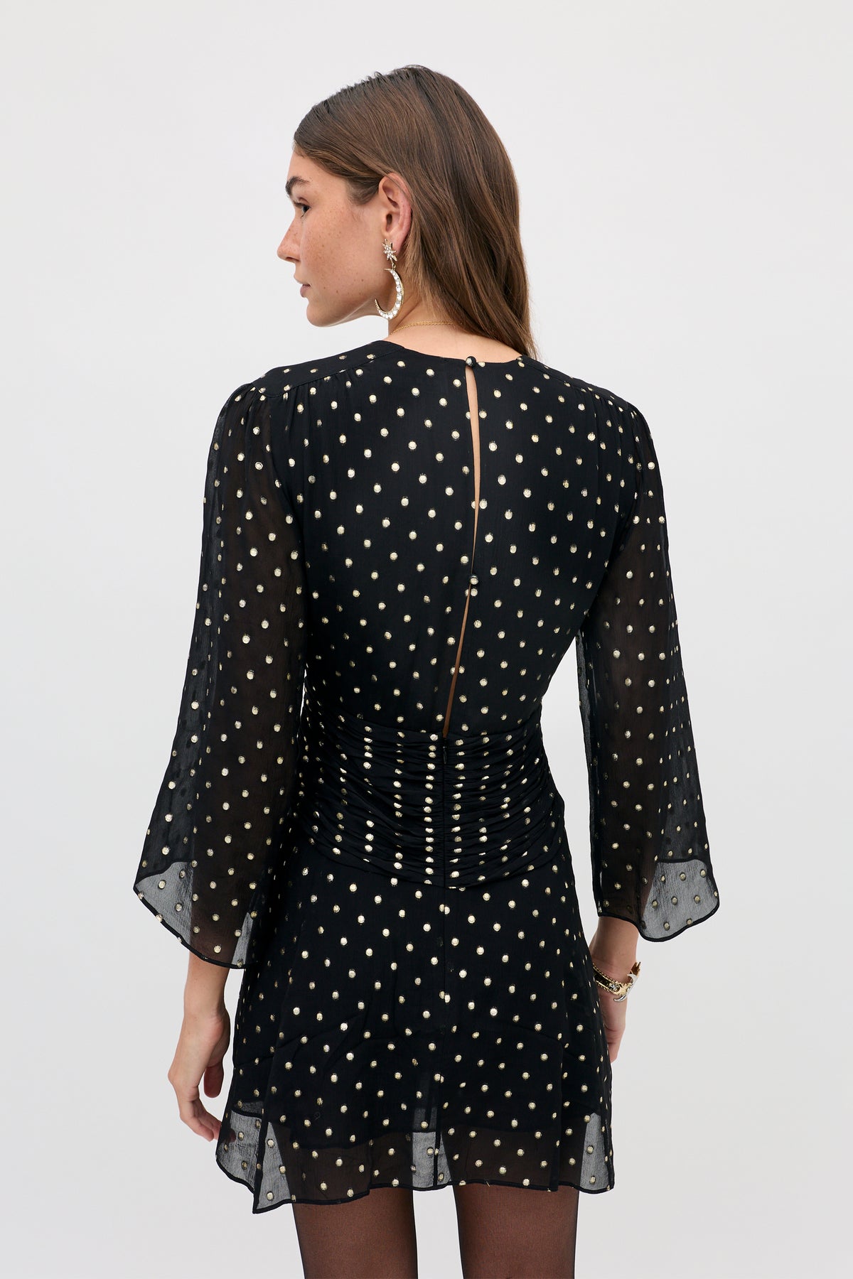 Back view of the Clove - Gold Spot Black mini dress, featuring black fabric with metallic gold polka dots, sheer three-quarter sleeves, and a ruched back detail.