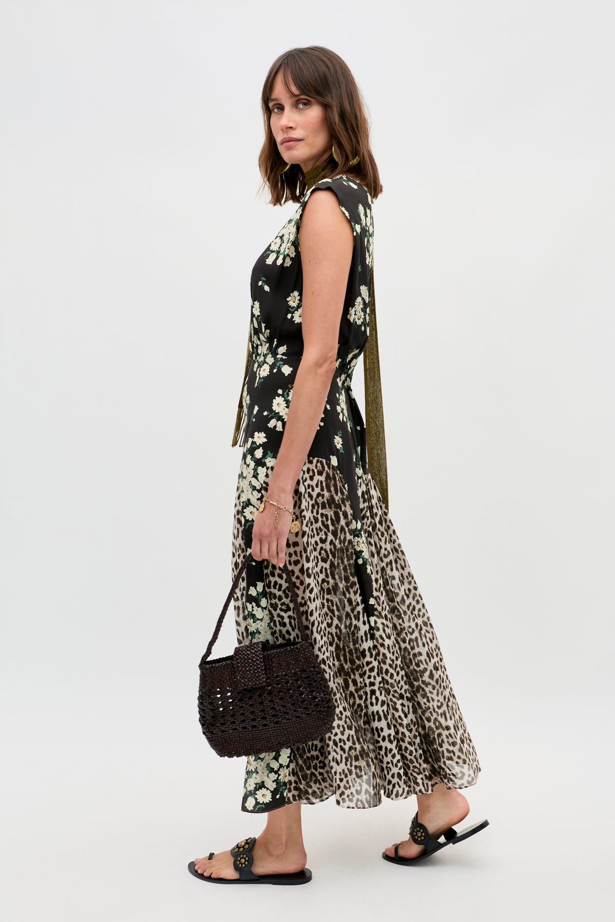 A woman stands against a plain white background wearing the Coralie - Amelia Gremlin Mix Black sleeveless floral and leopard print dress, black sandals, and holding a woven brown handbag.