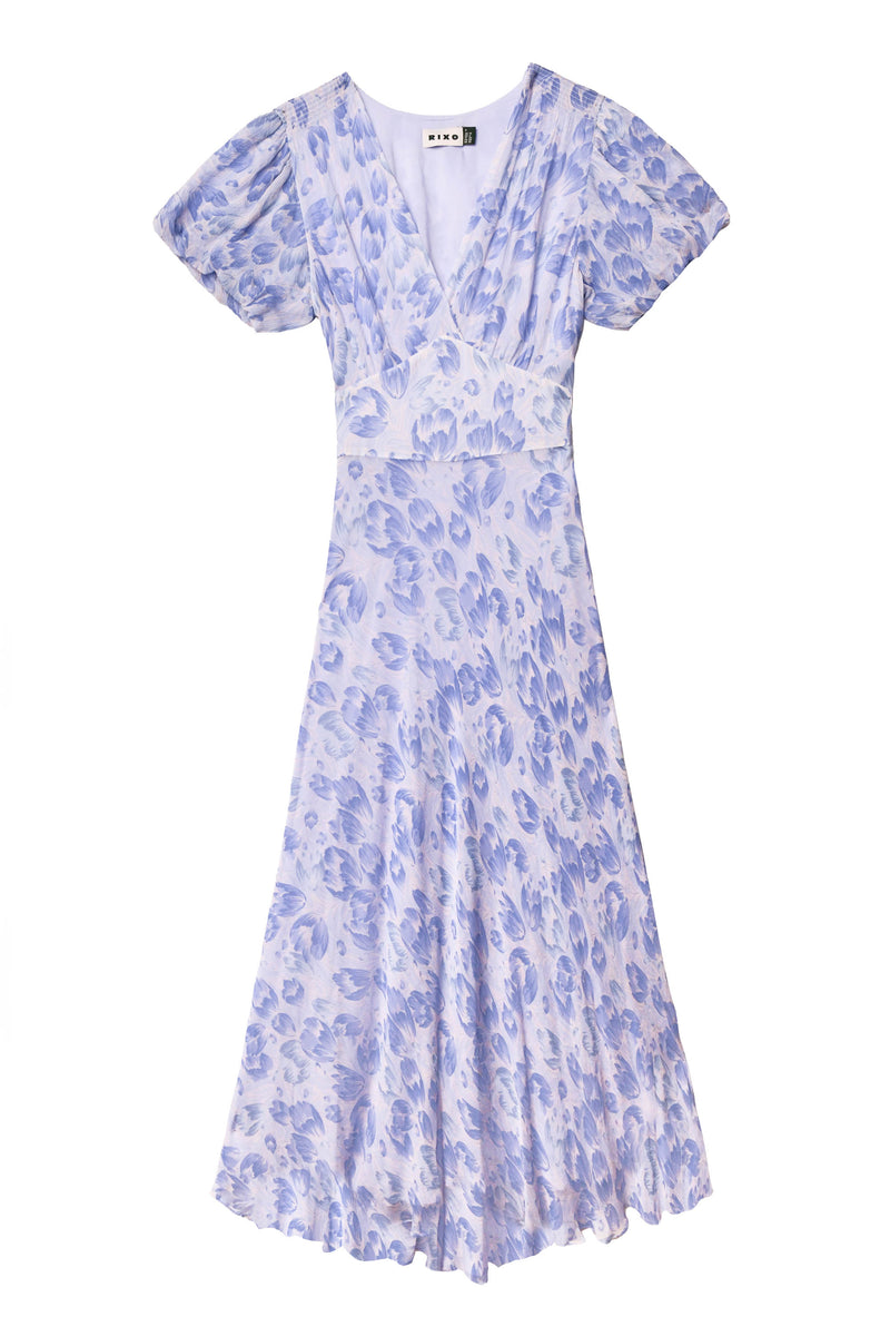 Perfect for summer events, the Corrine - Tulip Fields Cornflower Blue dress is a light purple midi with a v-neckline, short flutter sleeves, fitted waist, and gently flared skirt in a blue and lavender floral print.