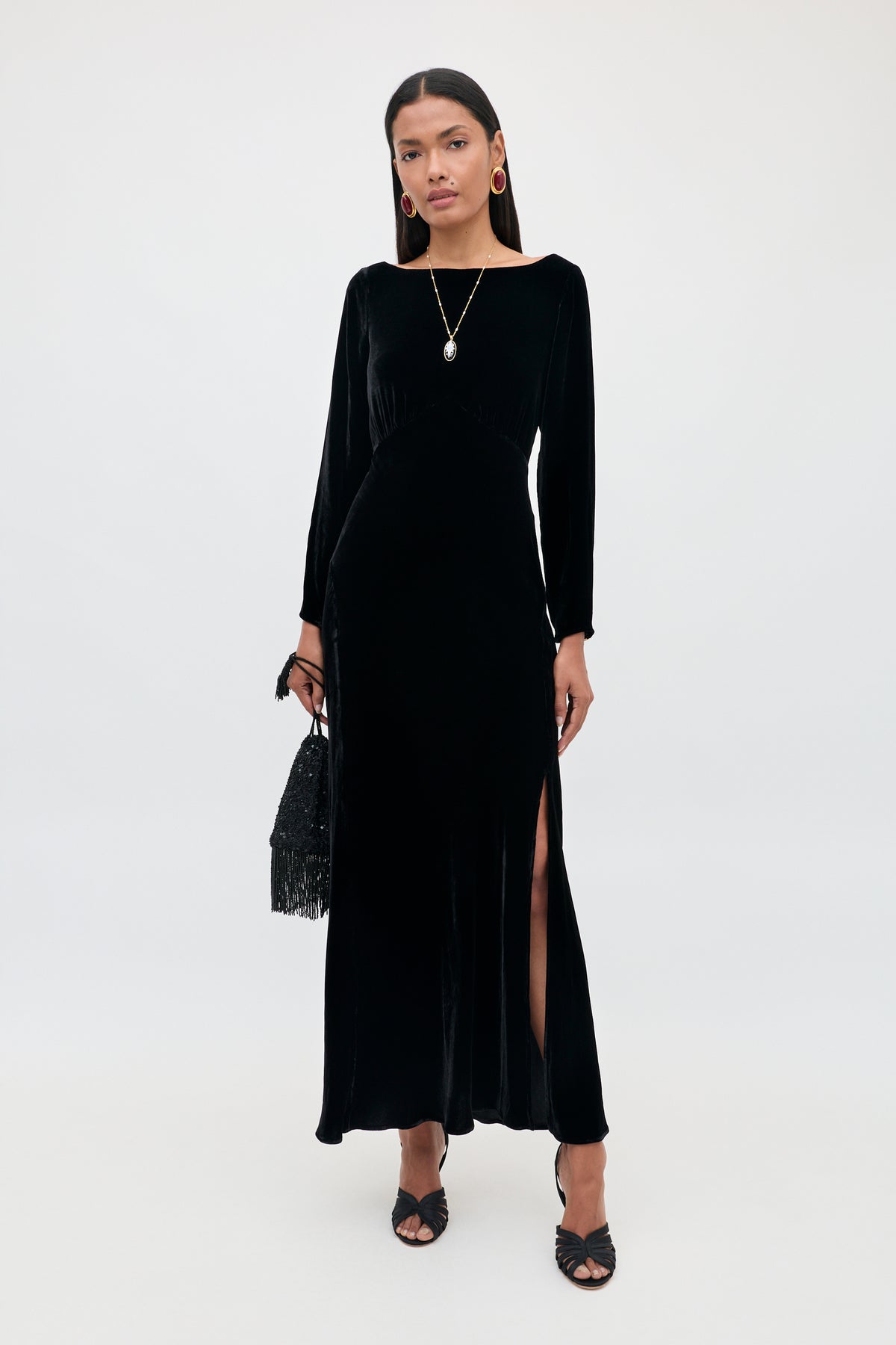 Wearing the Devina - Black gown, a woman stands against a plain background. The long velvet dress features long sleeves and a side slit, highlighting her sophisticated style. She completes the look with black heels, a black handbag, gold hoop earrings, and a pendant necklace.