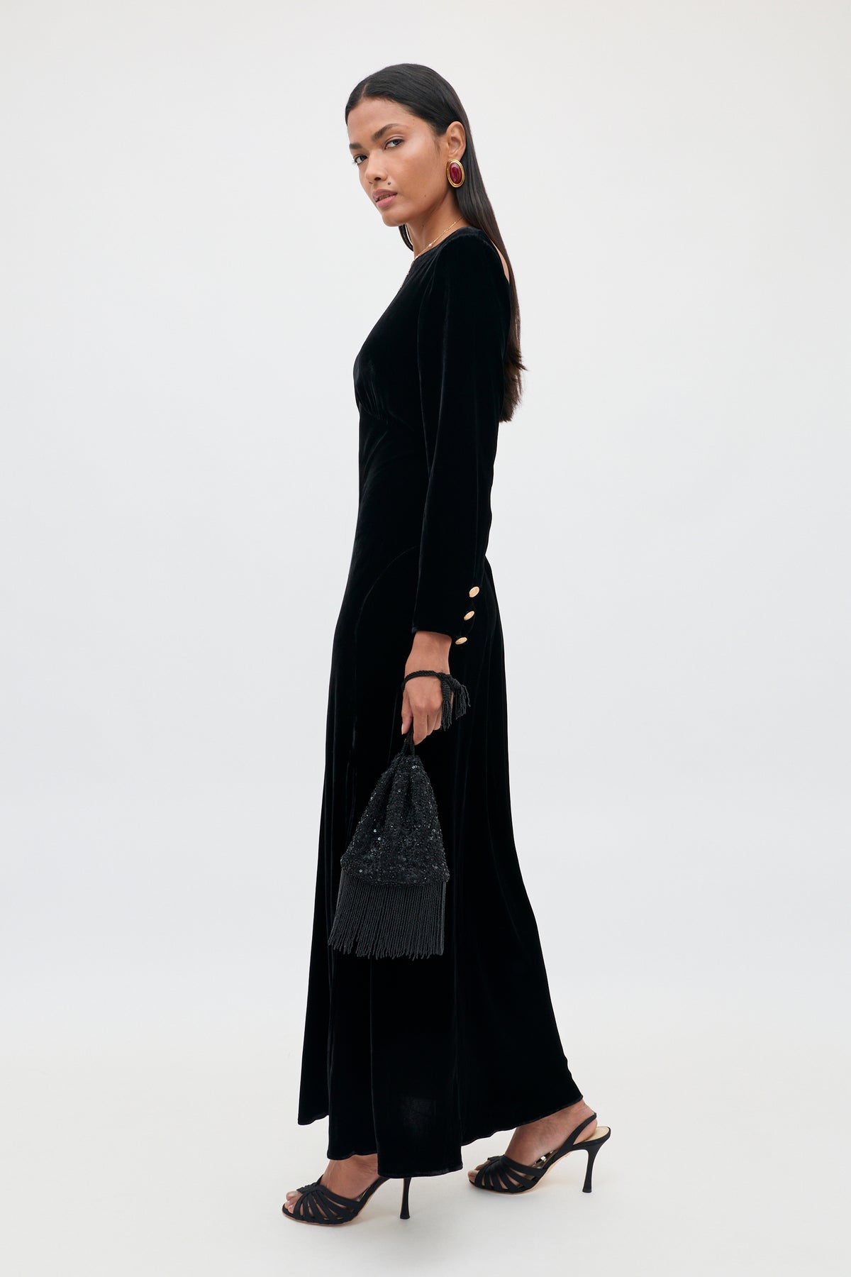 A woman stands sideways against a plain background, wearing the Devina – Black long black velvet gown with gold button details on the sleeves. She carries a black fringed handbag, wears black heels, and accessorizes with statement earrings. Her long dark hair complements the elegant look.