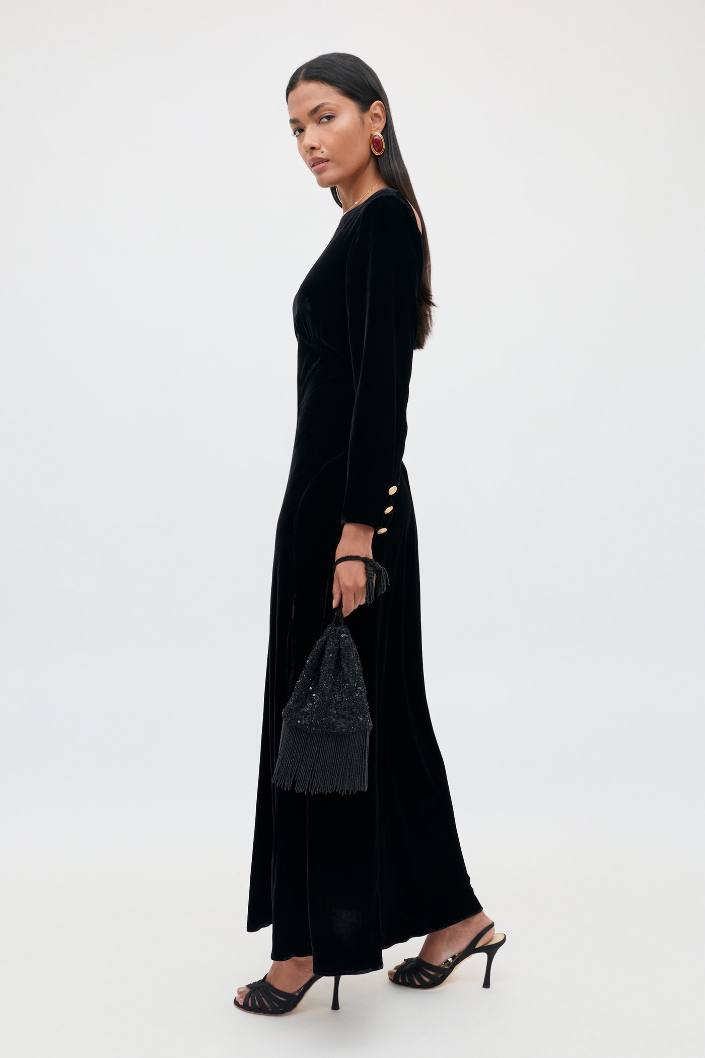 A woman stands sideways against a plain background, wearing the Devina – Black long black velvet gown with gold button details on the sleeves. She carries a black fringed handbag, wears black heels, and accessorizes with statement earrings. Her long dark hair complements the elegant look.