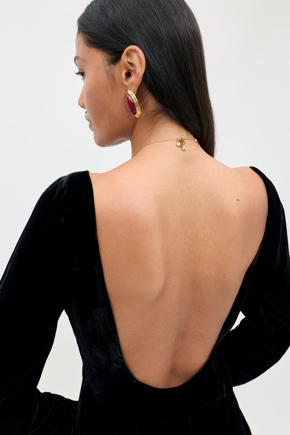 A woman with long dark hair is shown from behind against a plain, light background, wearing the Devina - Black gown—an elegant black velvet dress with a deep open back. She accessorizes with a gold necklace and large red and gold earrings.
