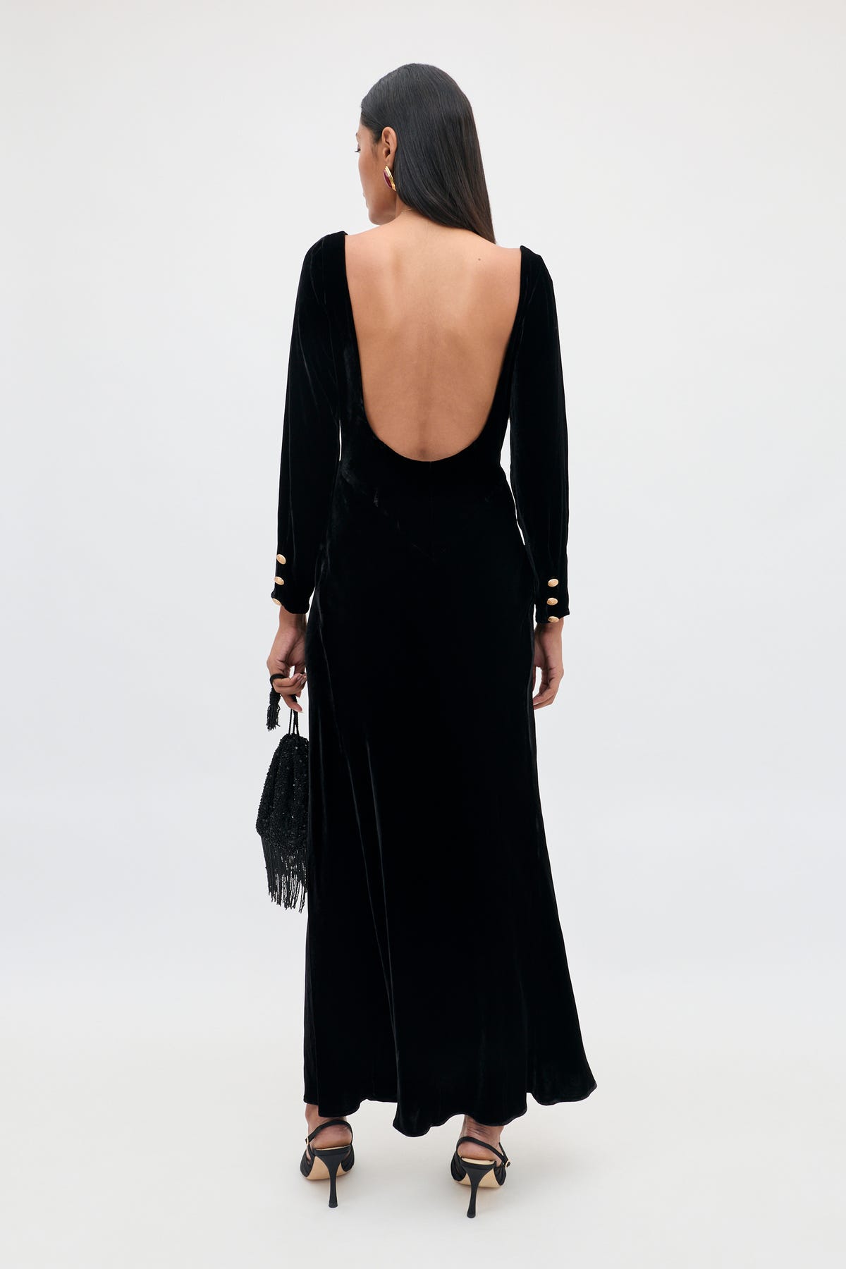 A woman stands facing away, perfectly showcasing the dramatic open back of the Devina – Black gown: a long, elegant evening dress crafted from black velvet with long sleeves and gold buttons. She completes the look with a black handbag and black high heels.