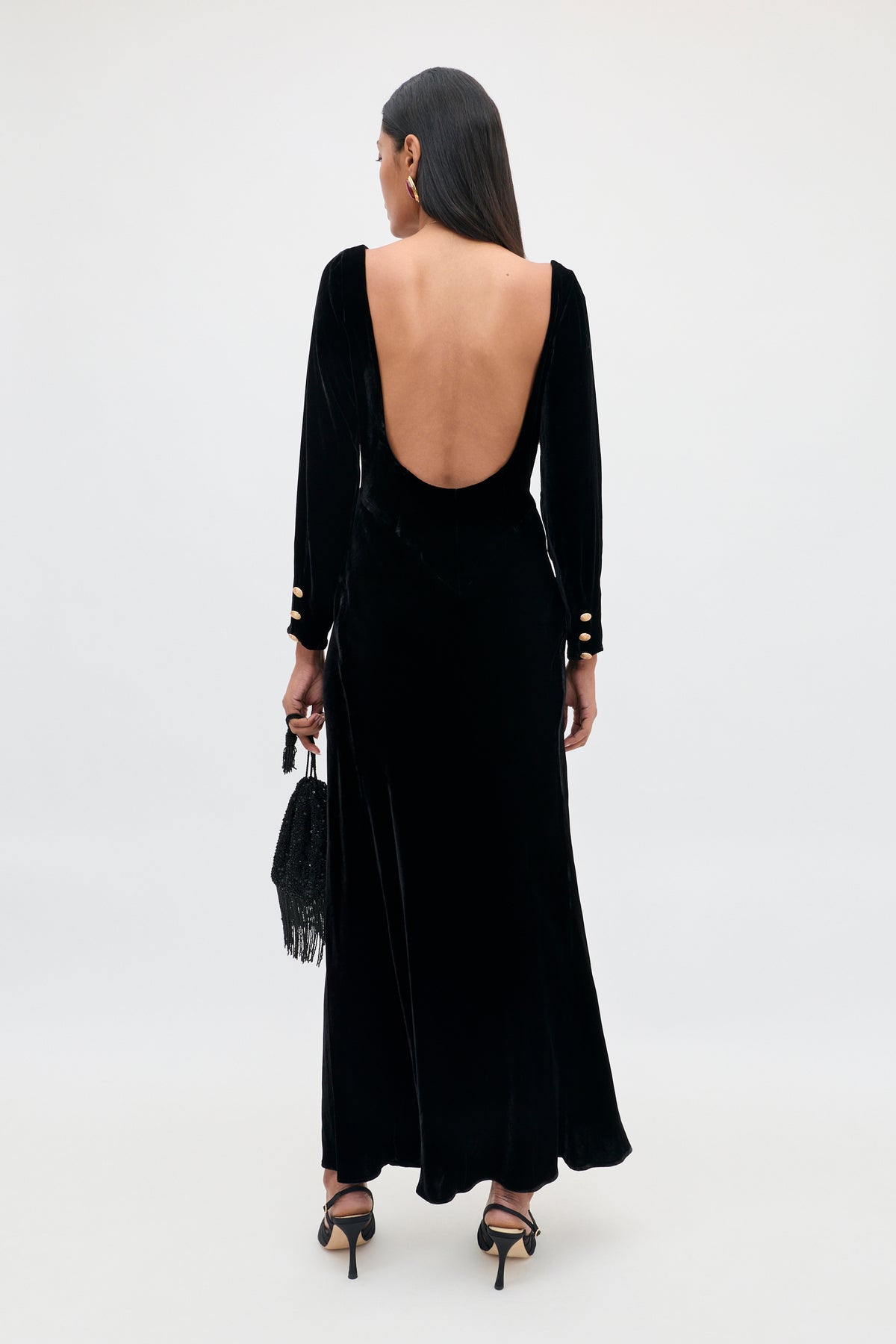 A woman stands facing away, perfectly showcasing the dramatic open back of the Devina – Black gown: a long, elegant evening dress crafted from black velvet with long sleeves and gold buttons. She completes the look with a black handbag and black high heels.