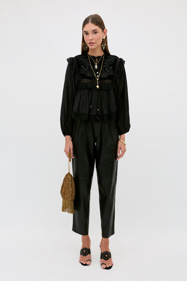 A woman stands against a plain background wearing the Diann - Lilly Embroidery Black long-sleeve blouse, paired with black pants, black open-toe heels, statement earrings, layered necklaces, and holding a gold beaded handbag with fringe.