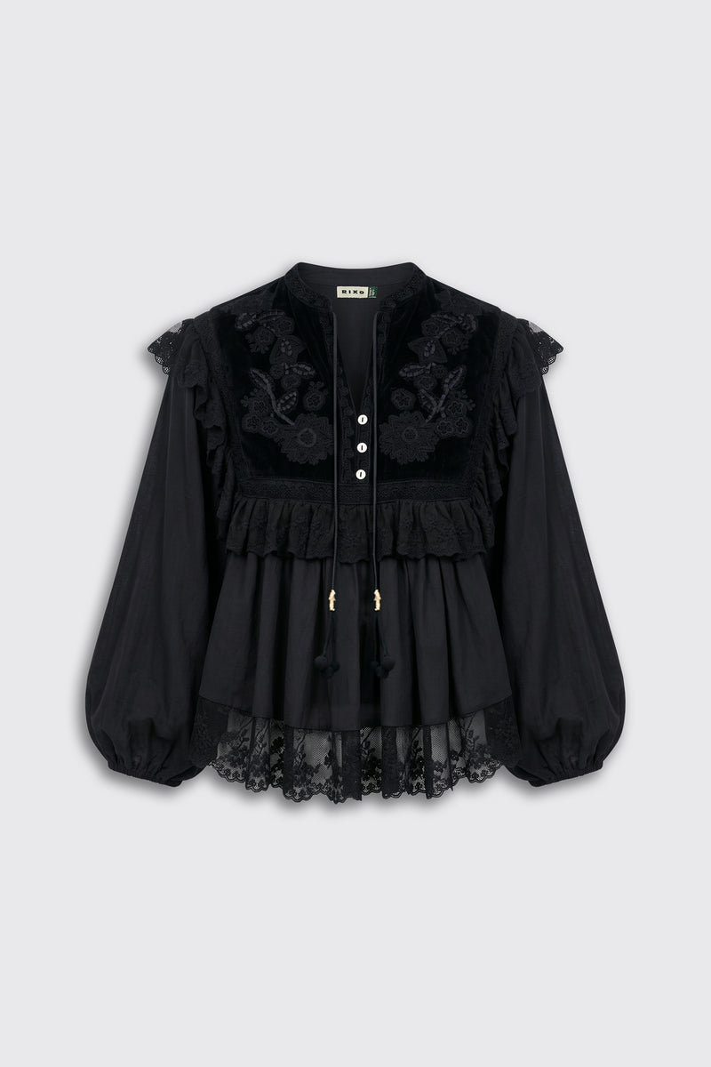 Diann - Lilly Embroidery Black is a black long-sleeve blouse featuring lace detailing, floral embroidery, ruffles, and a row of white buttons down the front. Designed for a loose fit, it has sheer lace edging at the sleeves and hem.