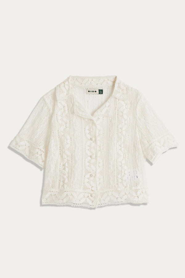 The Diona - Ivory is a short-sleeved, vintage-inspired lace blouse featuring a collared neckline, button front, intricate geometric and floral patterns, boxy shape, slightly cropped length, and a delicate airy look.