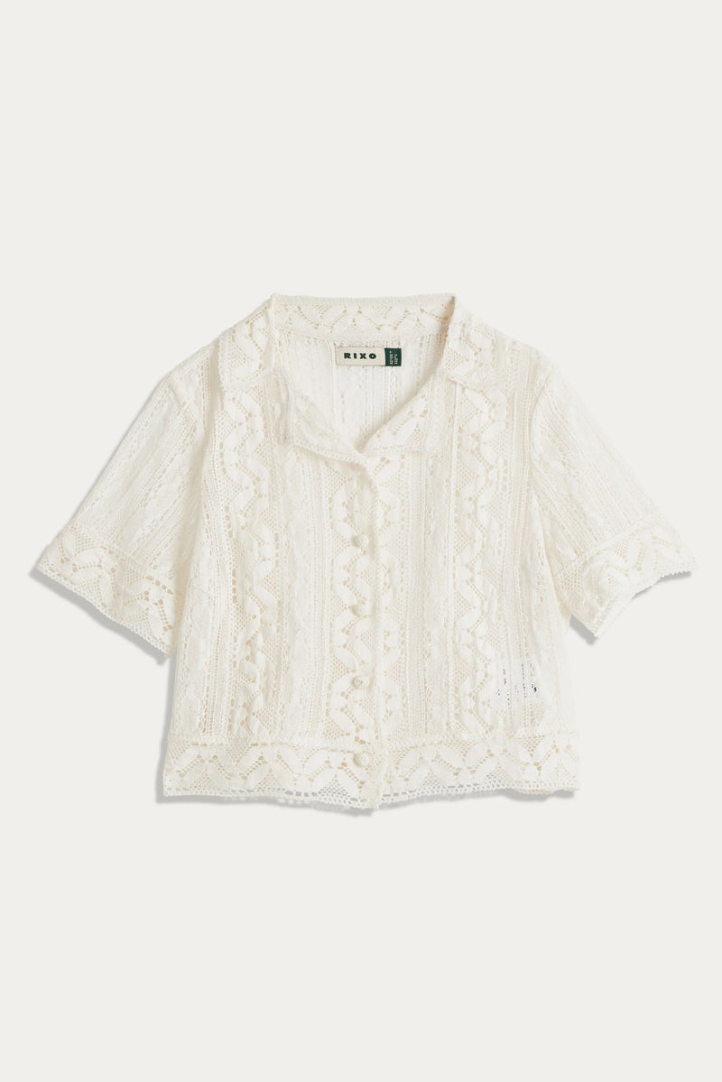 The Diona - Ivory is a short-sleeved, vintage-inspired lace blouse featuring a collared neckline, button front, intricate geometric and floral patterns, boxy shape, slightly cropped length, and a delicate airy look.