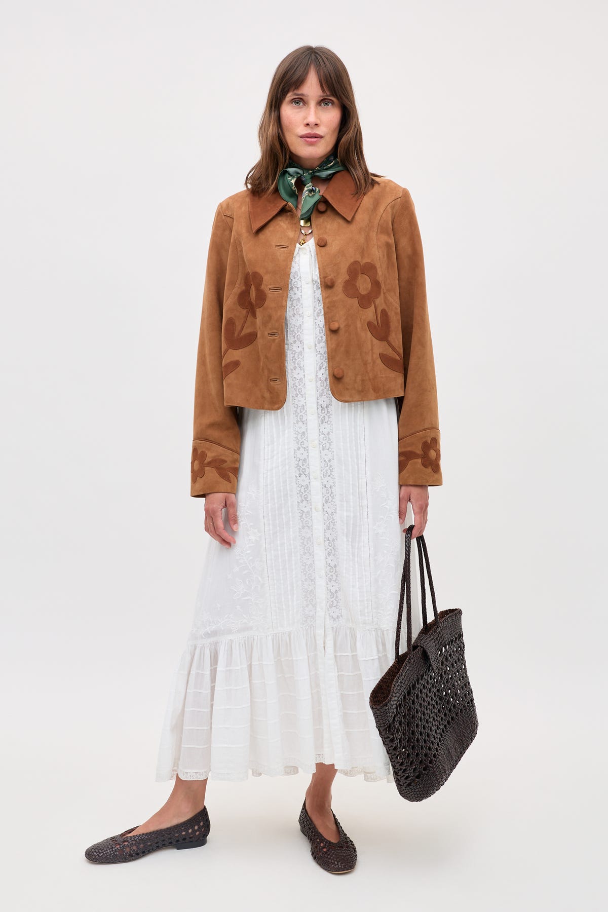 A woman stands against a plain background, wearing the Dionne - Retro Daisy brown suede jacket with daisy appliqué over a white lace dress, green scarf, brown woven shoes, and holding a black woven tote bag.