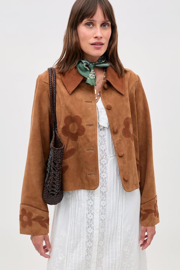 A woman in the Dionne - Retro Daisy brown suede jacket with daisy appliqué over a white lace dress, a green scarf, and holding a woven brown bag stands against a plain light background.