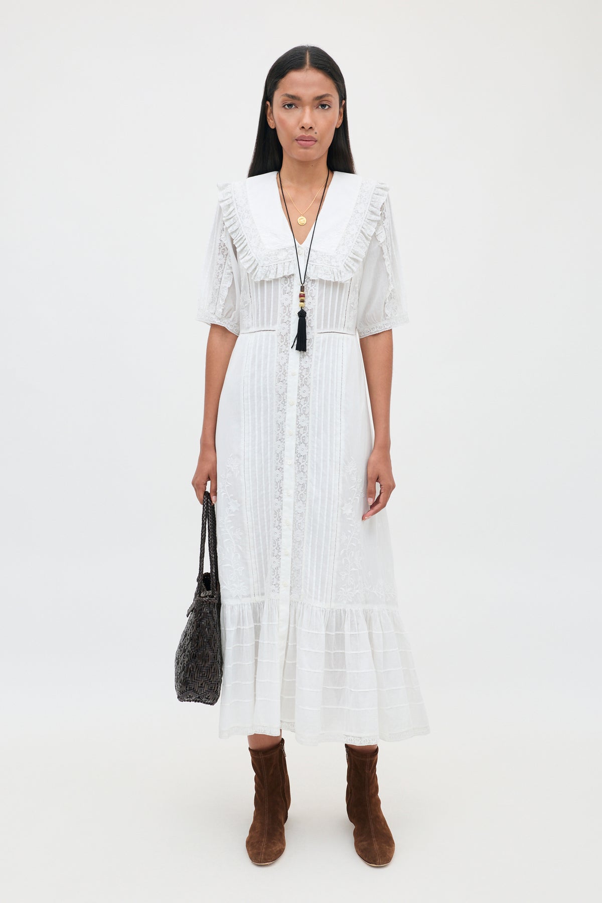 A woman stands against a white background wearing the Edina - Pansy Embroidery White dress, featuring delicate lace embroidery details, paired with brown boots, a black tasseled necklace, and holding a black handbag at her side.