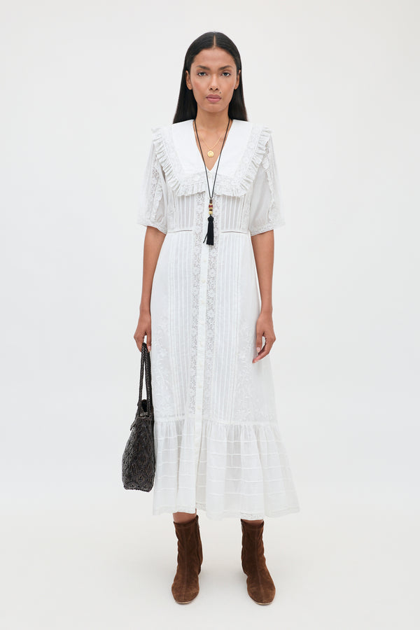 A woman stands against a white background wearing the Edina - Pansy Embroidery White dress, featuring delicate lace embroidery details, paired with brown boots, a black tasseled necklace, and holding a black handbag at her side.