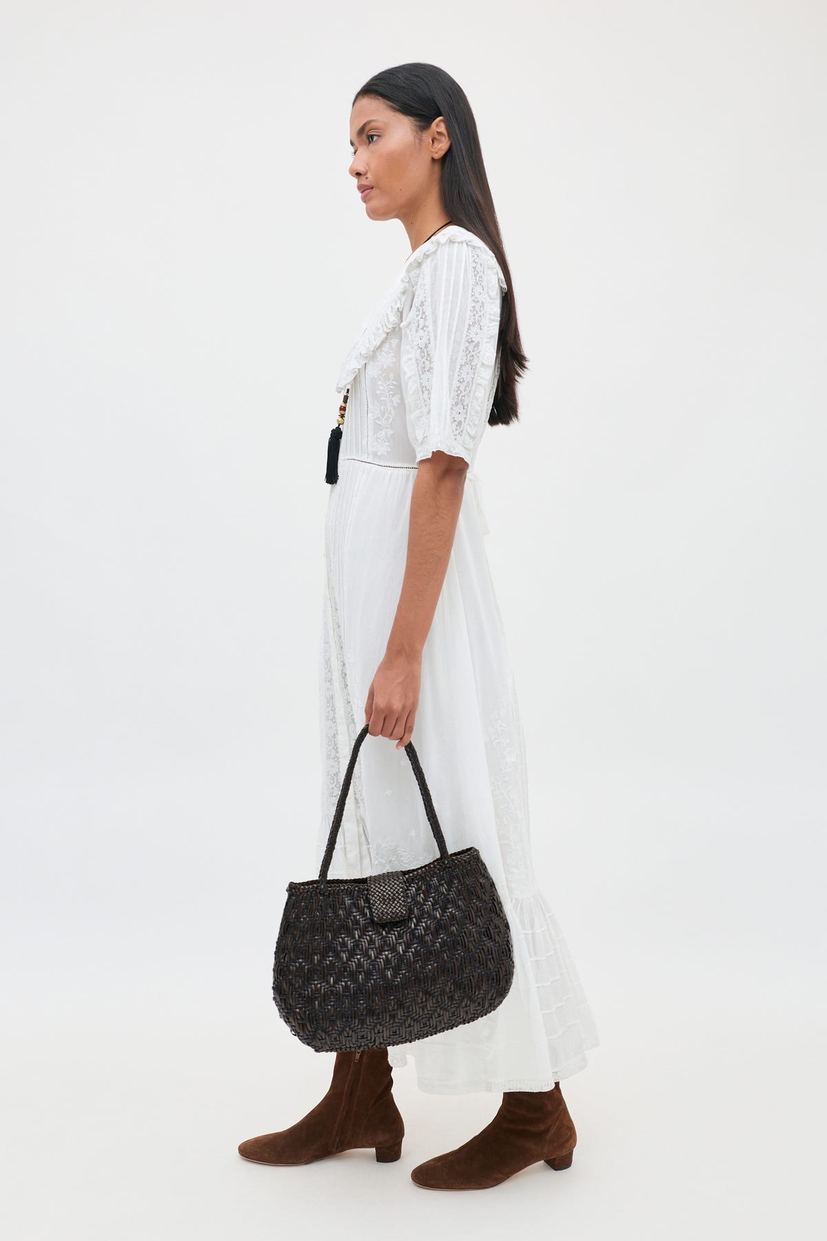 A woman stands in profile against a plain white background, wearing the Edina - Pansy Embroidery White dress—a knee-length white dress featuring delicate lace embroidery. She carries a black woven handbag, has long dark hair, and wears brown suede boots.