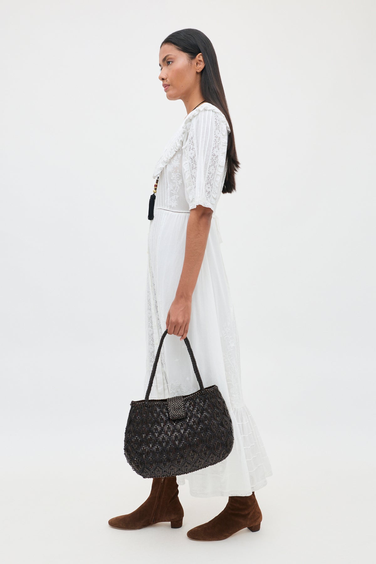 A woman stands in profile against a plain white background, wearing the Edina - Pansy Embroidery White dress—a knee-length white dress featuring delicate lace embroidery. She carries a black woven handbag, has long dark hair, and wears brown suede boots.