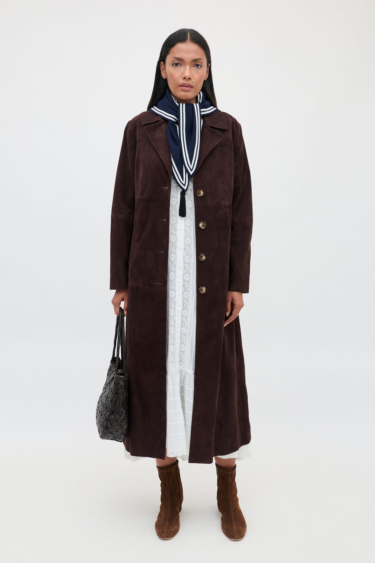 A woman wears the Edina - Pansy Embroidery White dress, layered with a long brown coat and styled with a navy and white scarf around her neck. She carries a patterned handbag and pairs the look with brown heeled ankle boots, standing against a simple light background.