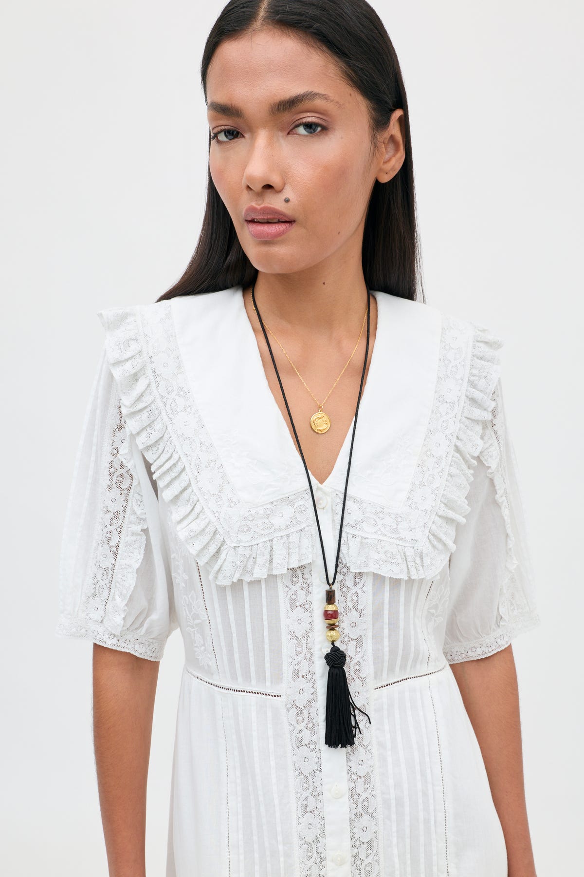 A woman with long dark hair stands against a plain white background, looking at the camera. She wears the Edina - Pansy Embroidery White dress—a white lace design featuring a ruffled collar and intricate lace embroidery—styled with two layered necklaces: one adorned with a gold coin pendant and the other with a black tassel.