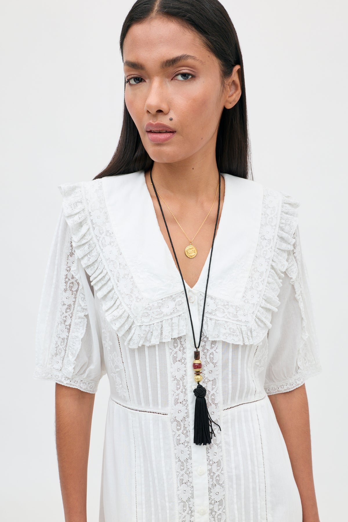 A woman with long dark hair stands against a plain white background, looking at the camera. She wears the Edina - Pansy Embroidery White dress—a white lace design featuring a ruffled collar and intricate lace embroidery—styled with two layered necklaces: one adorned with a gold coin pendant and the other with a black tassel.