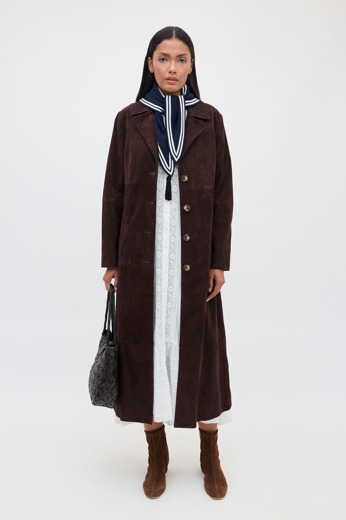 Wearing the Edina - Pansy Embroidery White dress, a woman styles her look with a long brown suede coat, a blue and white scarf, brown boots, and carries a textured black bag while standing against a plain light background.