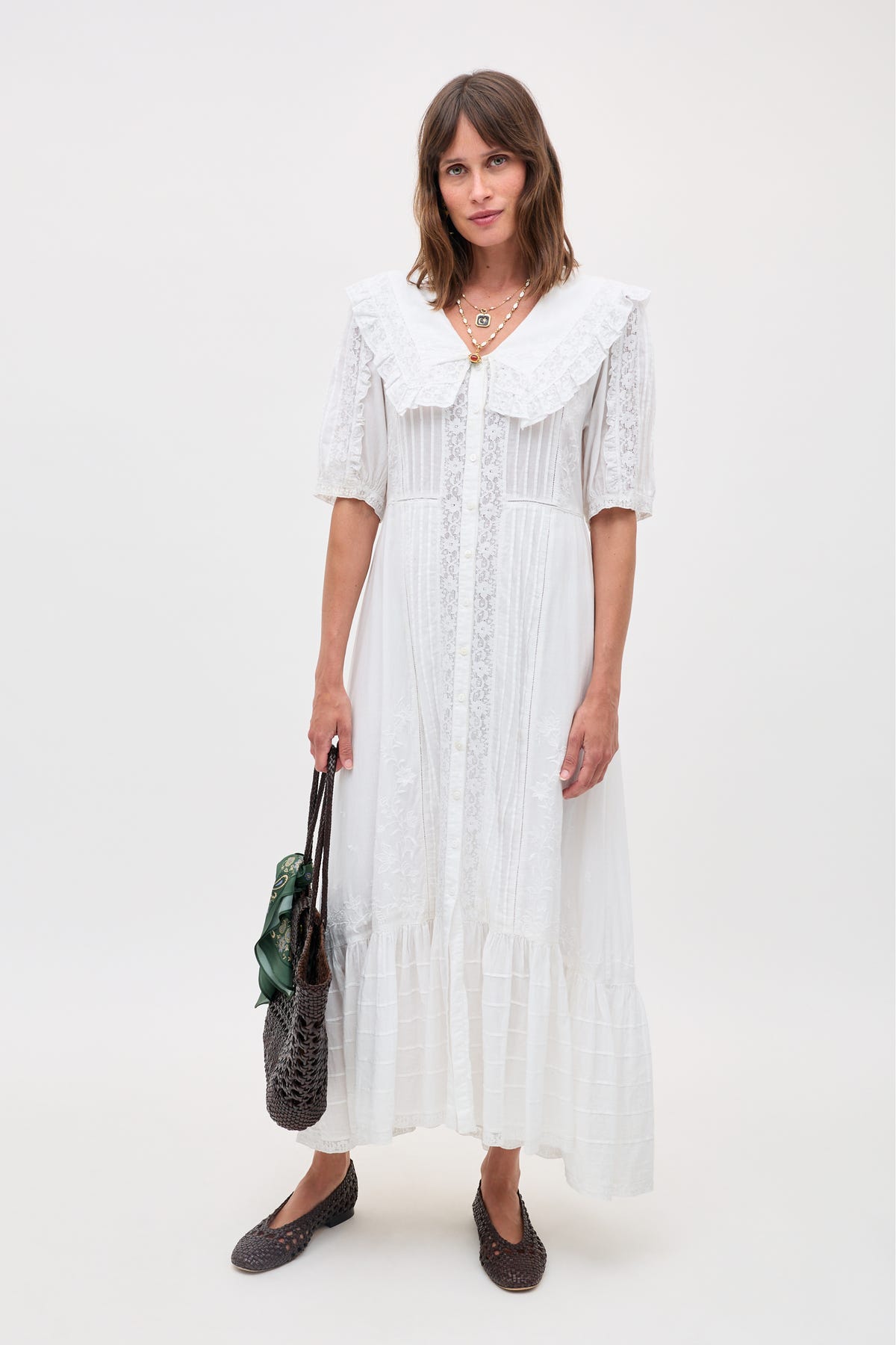 A woman wears the Edina - Pansy Embroidery White long lace blouse dress with puffed sleeves and a large collar, standing against a plain background, holding two bags and wearing brown woven flats.
