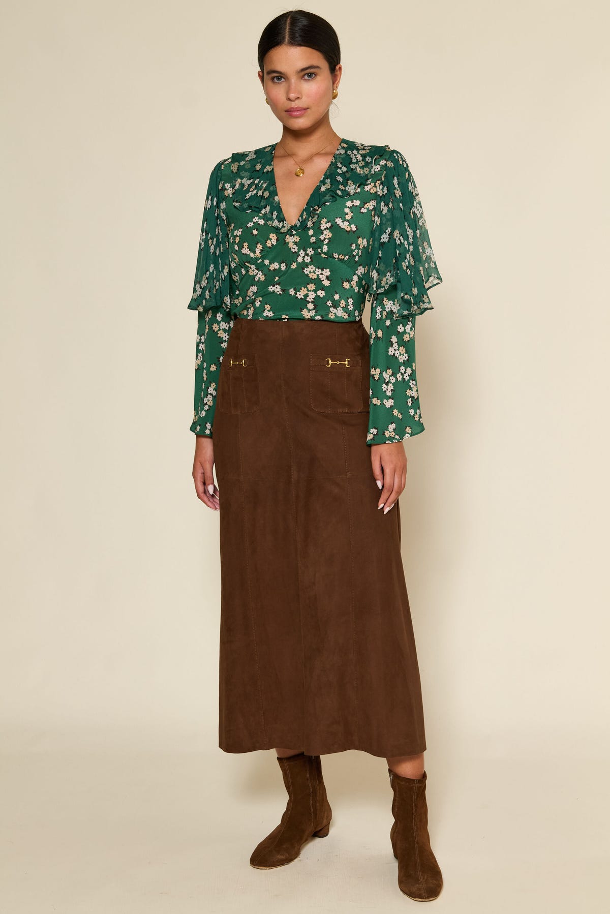 A woman with dark hair pulled back and a neutral expression stands against a plain background wearing a green floral blouse with ruffled sleeves, a high-waisted A-line vintage-inspired skirt with front pockets, and Edwina - Brown suede boots.