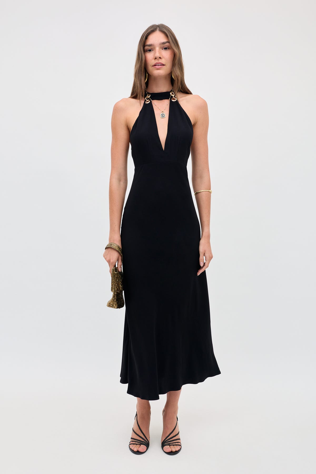 A woman stands against a plain background wearing the Eleanora - Black halter-neck dress with gold accents, paired with a small gold purse, black strappy heels, gold bracelets, and her long hair styled loose.