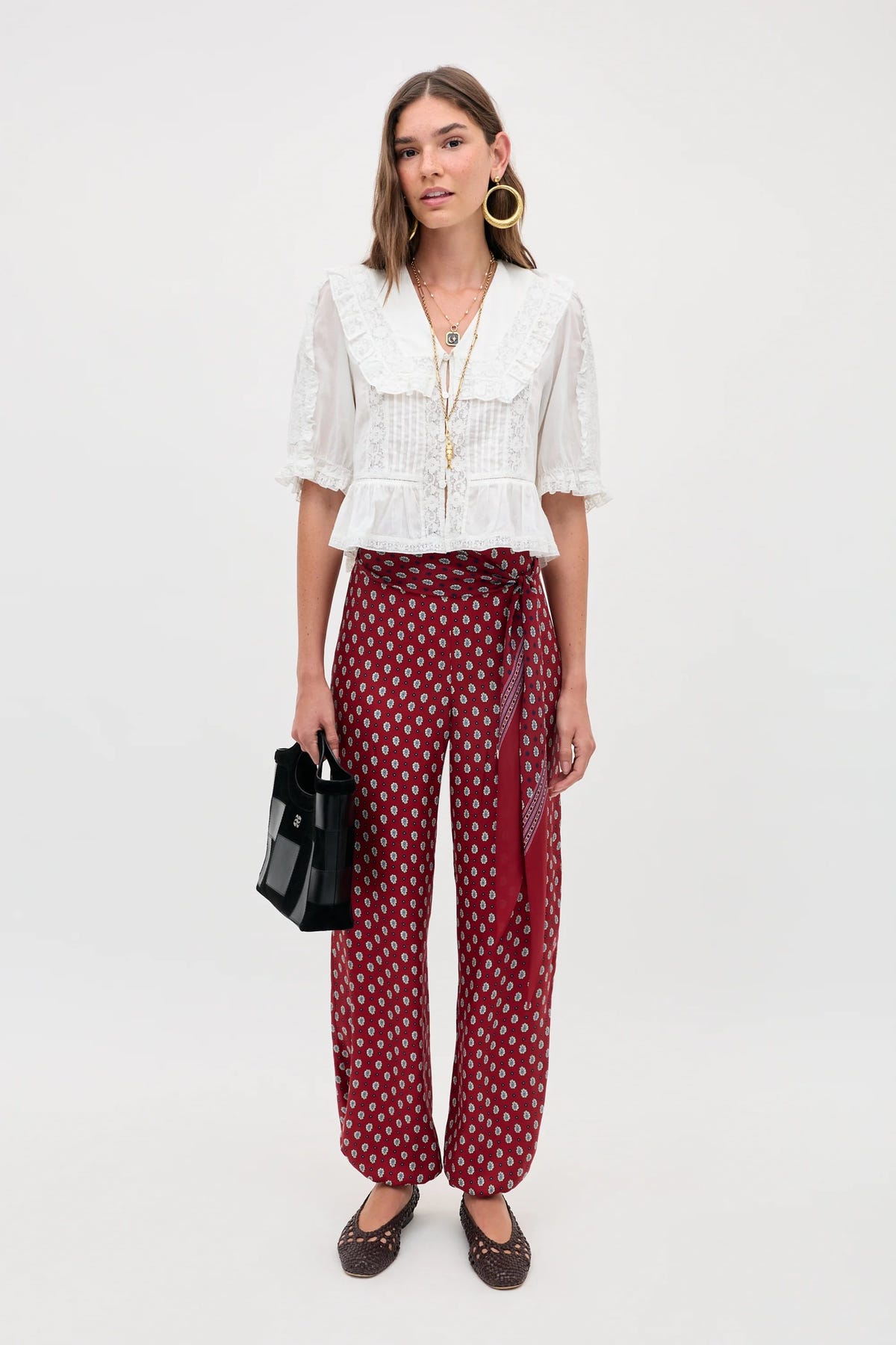 A woman stands against a plain background wearing the Ellery - White blouse with a ruffled collar, styled with red patterned wide-leg pants, brown shoes, gold hoops, layered necklaces, and holding a black handbag.