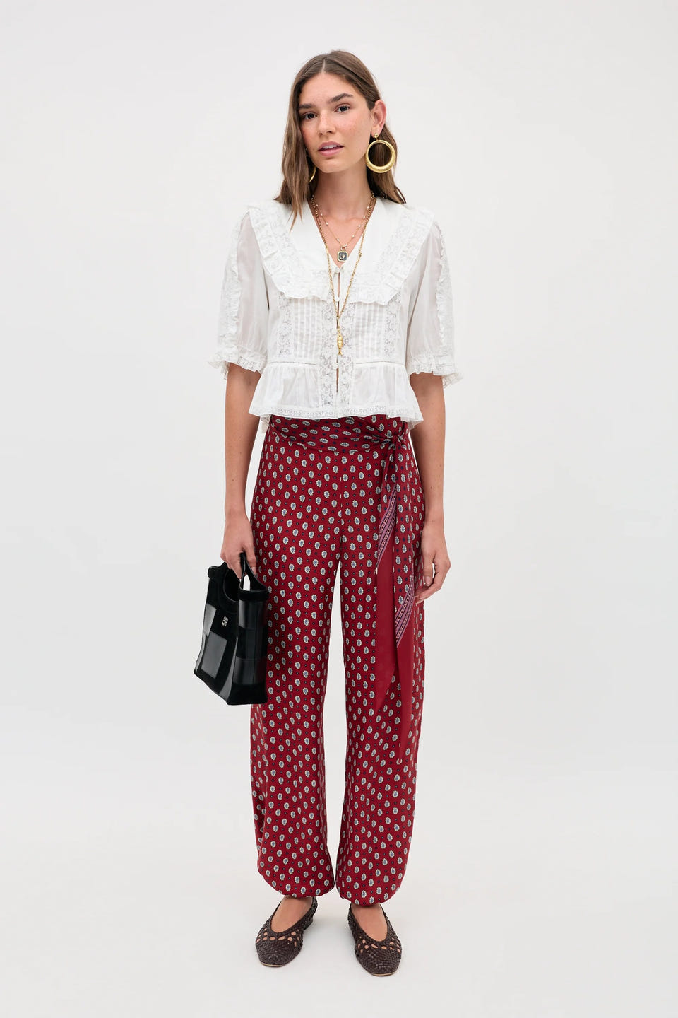 A woman stands against a plain background wearing the Ellery - White blouse with a ruffled collar, styled with red patterned wide-leg pants, brown shoes, gold hoops, layered necklaces, and holding a black handbag.