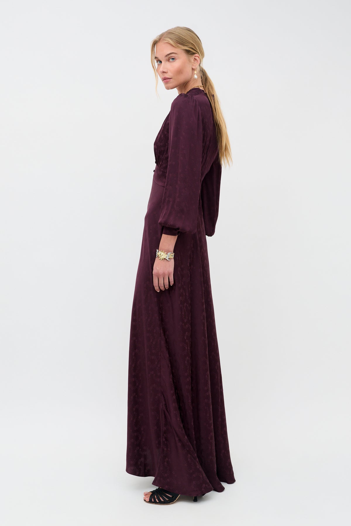 Here is the revised sentence using the product data: Woman wearing the Emory - Floral Trail Jacquard Wine, a long-sleeved, floor-length dress in a rich wine hue with a subtle floral pattern. She stands sideways and looks at the camera, her long blonde hair styled in a low ponytail, and she pairs the look with black sandals.
