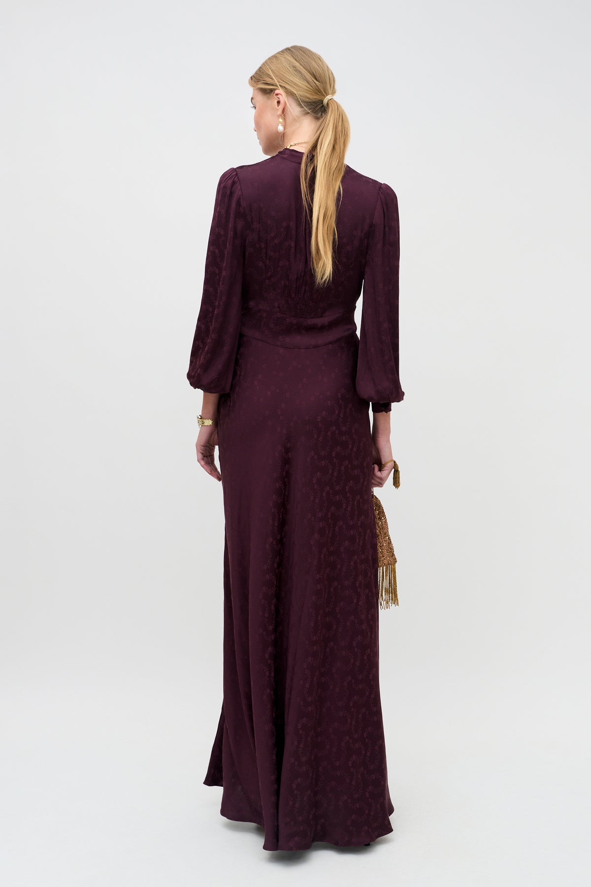 A woman with long blonde hair in a ponytail stands with her back to the camera, wearing the Emory - Floral Trail Jacquard Wine dress—a long-sleeved, floor-length burgundy gown—and holding a small gold fringe bag.