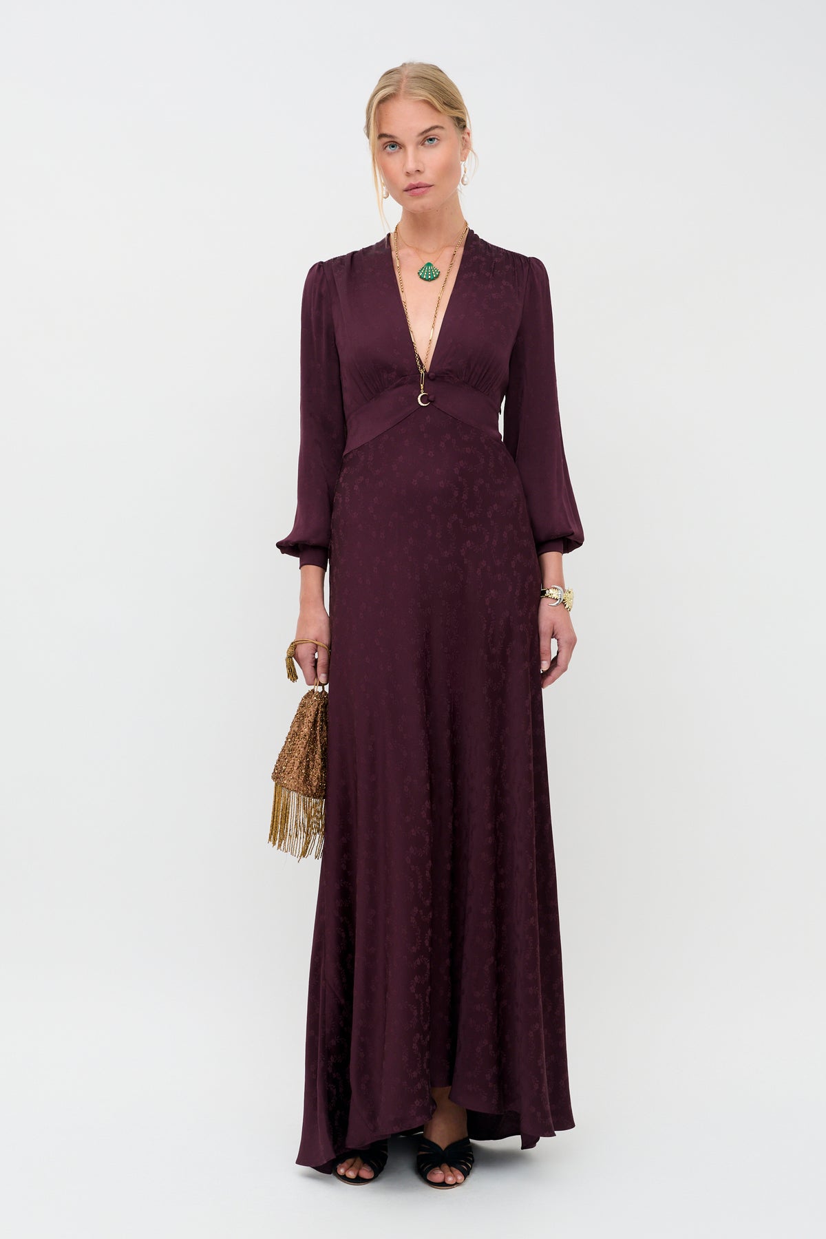 A woman stands against a plain white background wearing the Emory - Floral Trail Jacquard Wine dress, a long, deep burgundy gown with long sleeves. She holds a brown fringed handbag and accessorizes with a necklace and bracelets.