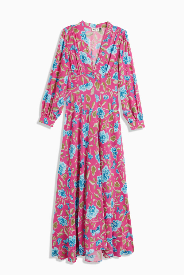The Emory - Butterfly Floral Magenta is a long-sleeved, floor-length dress in pink with a deep V-neckline and a bold floral print of blue flowers and green leaves.