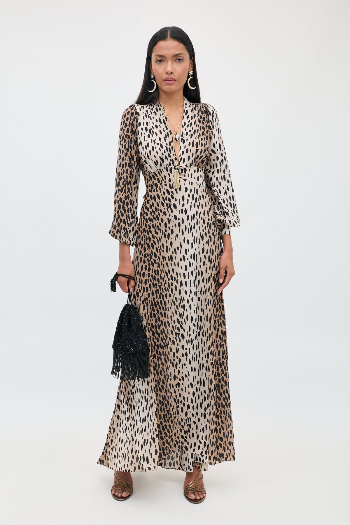 A woman stands against a plain background wearing the Emory - Antelope Spot Black, an elegant long dress crafted in luxurious silk crepe de chine with a deep V-neck. She accessorizes with hoop earrings, black sandals, and a small black fringe bag—perfect for any special occasion.