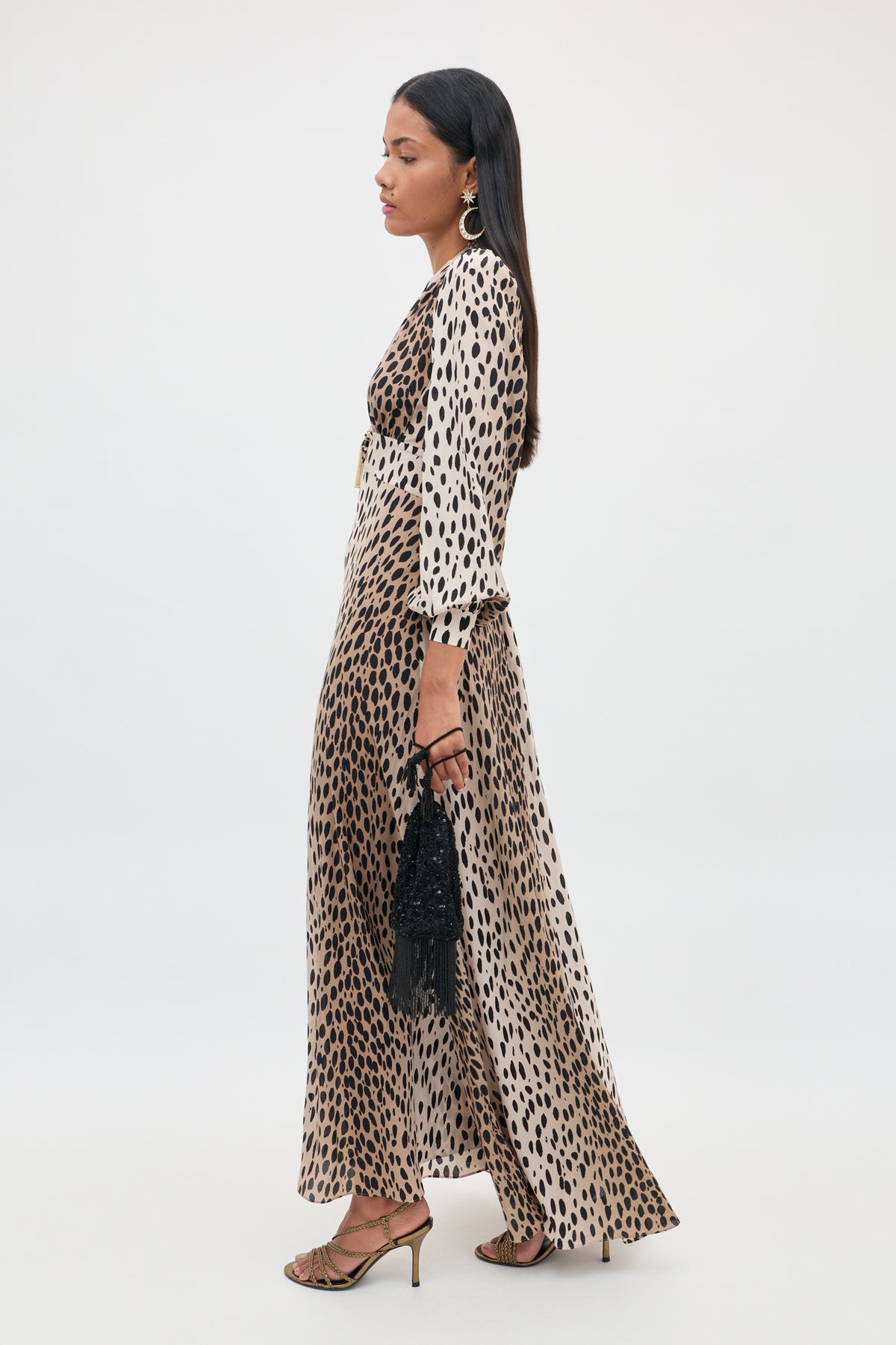 A woman standing in profile, wearing the Emory - Antelope Spot Black dress crafted from luxurious silk crepe de chine in an eye-catching antelope spot print. She styles it with long sleeves, strappy heels, hoop earrings, and carries a small black beaded bag against a plain white background—an effortlessly elegant look for special occasions.