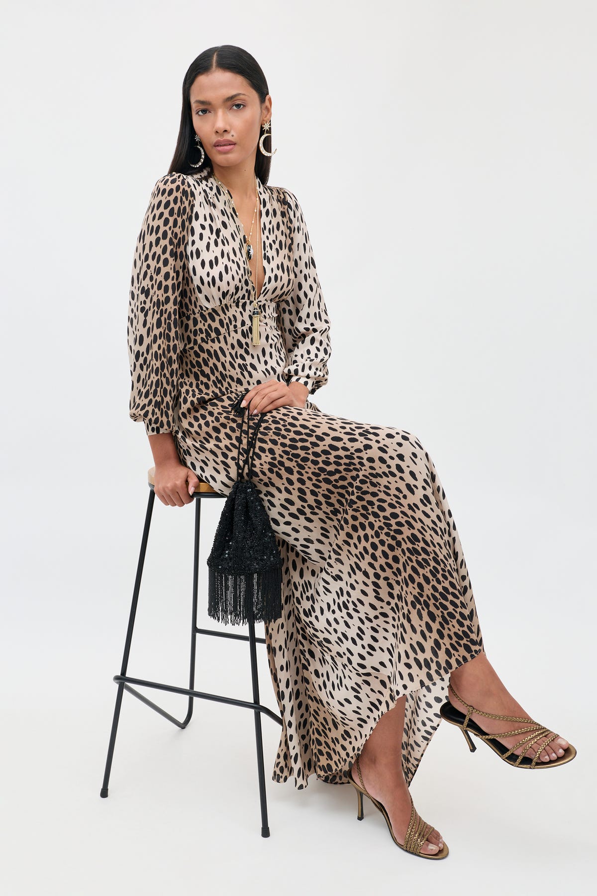 Seated gracefully on a tall stool, a woman wears the Emory - Antelope Spot Black dress, an elegant leopard-print design in luxurious silk crepe de chine with a plunging neckline. She completes her special occasion ensemble with gold sandals and statement earrings, holding a black fringed handbag. Her long, straight hair flows beautifully as she poses against a simple light backdrop.