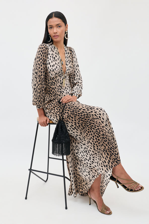 Seated gracefully on a tall stool, a woman wears the Emory - Antelope Spot Black dress, an elegant leopard-print design in luxurious silk crepe de chine with a plunging neckline. She completes her special occasion ensemble with gold sandals and statement earrings, holding a black fringed handbag. Her long, straight hair flows beautifully as she poses against a simple light backdrop.