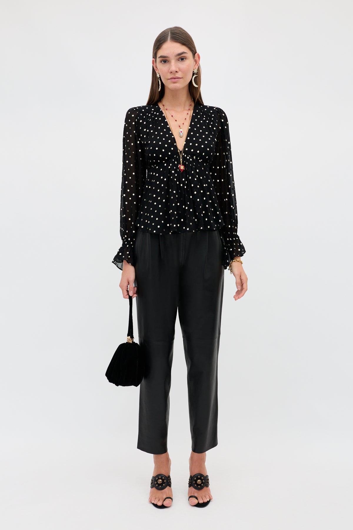 A woman wearing an Ethel - Gold Spot Black long-sleeved top with gold polka dots and black trousers, holding a small black handbag and wearing black heeled sandals.