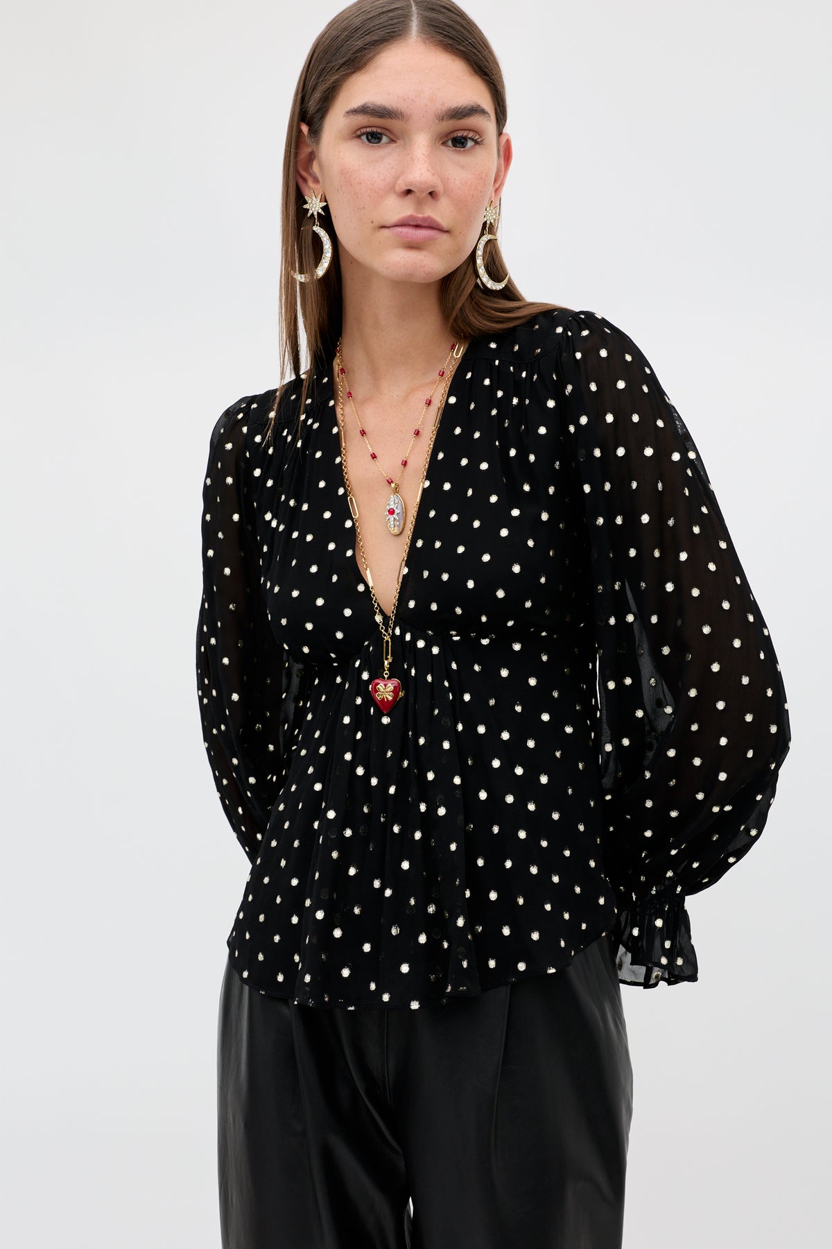 A woman wearing the Ethel - Gold Spot Black blouse, a black long-sleeved top with a gold polka-dot pattern and deep V-neck, styled with layered pendant necklaces and large star and moon earrings.