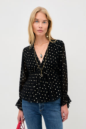 A woman wearing an Ethel - Gold Spot Black long-sleeved V-neck top with gold polka dots and ruffled cuffs, paired with blue jeans.