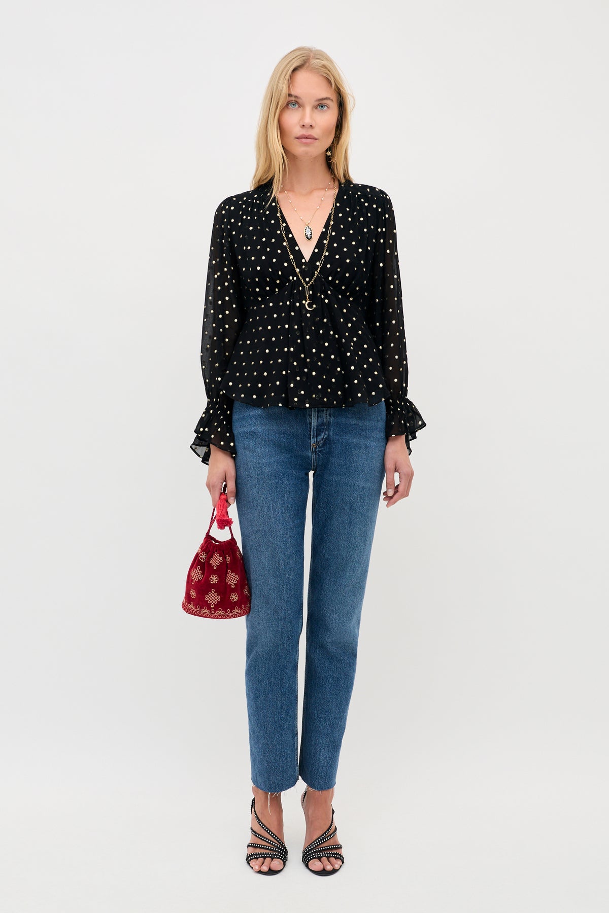 A woman wears an Ethel - Gold Spot Black long-sleeved blouse, blue jeans, and strappy black heels, holding a small red embellished drawstring bag.