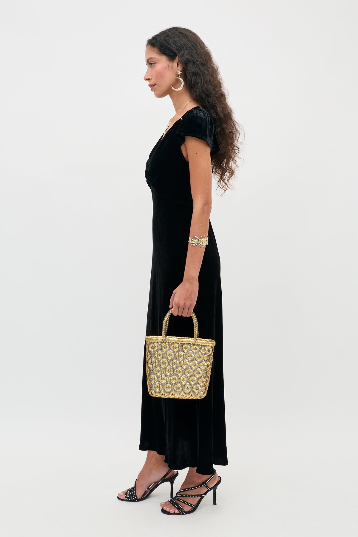 A woman wears an Ezra - Black maxi dress and embellished strappy heels while carrying a metallic gold woven basket handbag.