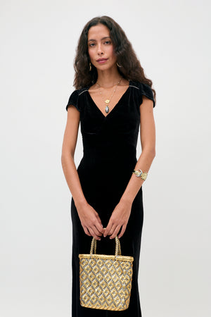 A person wearing the Ezra - Black short-sleeved velvet v-neck dress, layered gold necklaces, and a gold cuff bracelet, holding a metallic gold woven handbag.