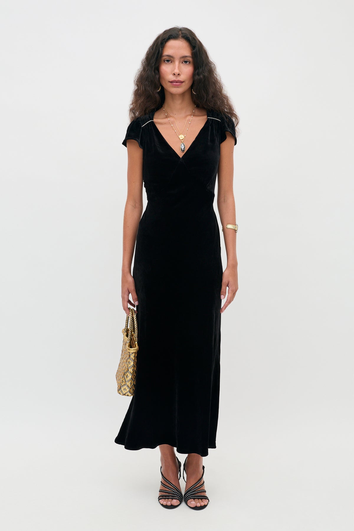 A person wearing the Ezra - Black midi dress with cap sleeves, holding a gold metallic handbag and wearing embellished strappy heels.
