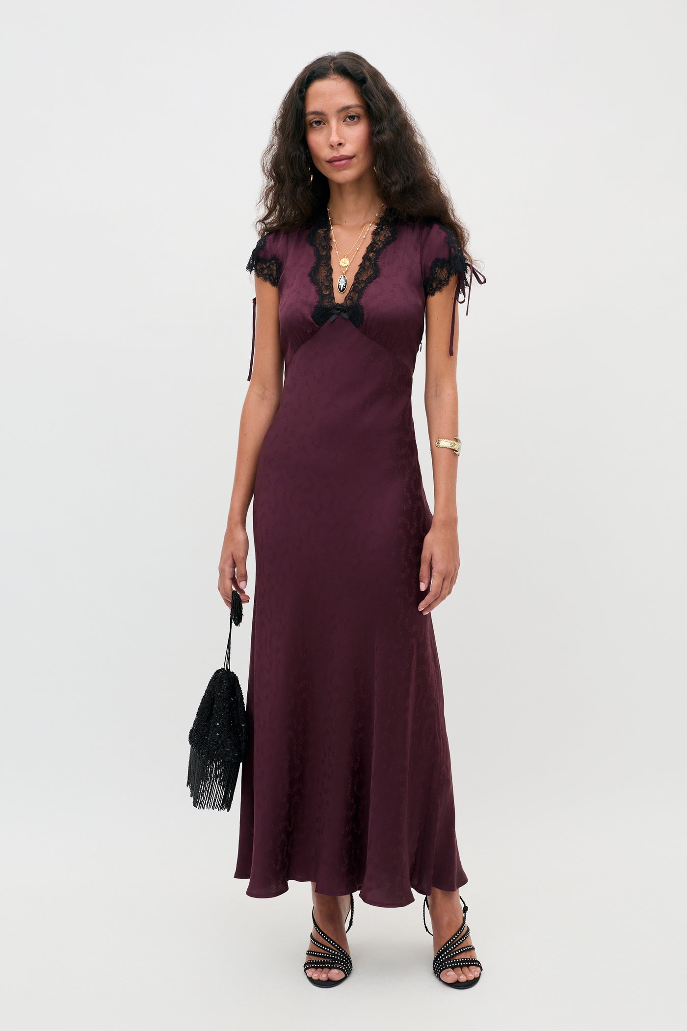 A woman stands against a plain background wearing the Fern - Floral Trail Jacquard Wine dress, a long, deep purple gown with black lace trim. She accessorizes with a black fringed bag, layered necklaces, and black strappy heels, her long curly brown hair cascading over her shoulders.