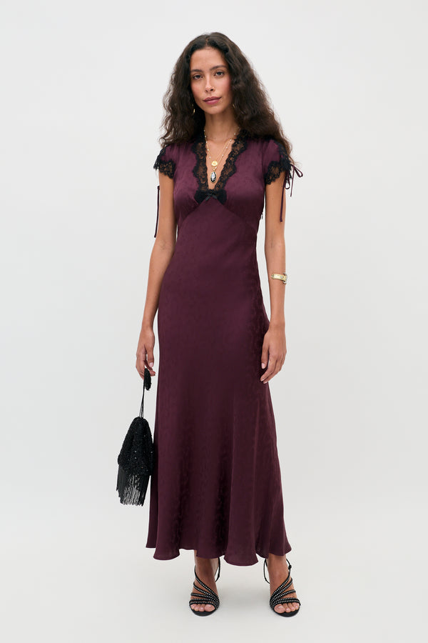 A woman stands against a plain background wearing the Fern - Floral Trail Jacquard Wine dress, a long, deep purple gown with black lace trim. She accessorizes with a black fringed bag, layered necklaces, and black strappy heels, her long curly brown hair cascading over her shoulders.