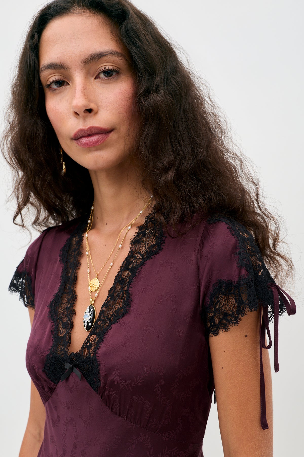 Woman with long wavy brown hair wearing the Fern - Floral Trail Jacquard Wine dress, styled with layered gold and black necklaces, standing against a plain light background.