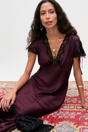 A woman in the Fern - Floral Trail Jacquard Wine dress with lace trim sits on a colorful patterned rug, wearing gold jewelry and holding a black beaded bag. She has long wavy hair and a confident expression.