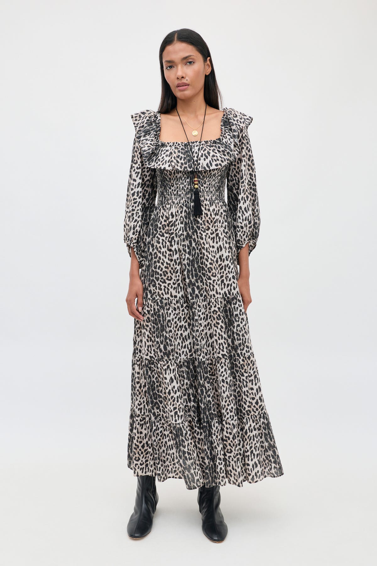 A person stands against a plain background wearing the Gabriel - Nina Leopard Mocha dress with ruffled sleeves and black ankle boots. They have long dark hair and are accessorized with layered necklaces.