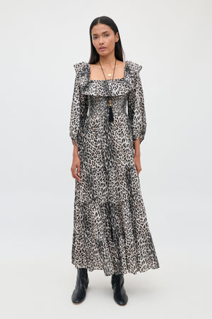 A person stands against a plain background wearing the Gabriel - Nina Leopard Mocha dress with ruffled sleeves and black ankle boots. They have long dark hair and are accessorized with layered necklaces.